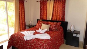 Een net opgemaakt bed met een rode bedspreide, twee kussens en een witte handdoek. Er is een nachtkastje met een lamp en een klein lade. De kamer heeft een raam met gordijnen, een airconditioning en een plafondventilator.