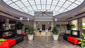 A spacious lobby with a high ceiling and a glass dome, featuring a reception desk, seating areas, and potted plants.