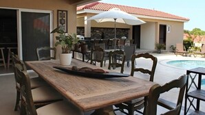 Un espacio de comedor al aire libre con una mesa y sillas de madera, rodeado de plantas en macetas y un paraguas blanco.