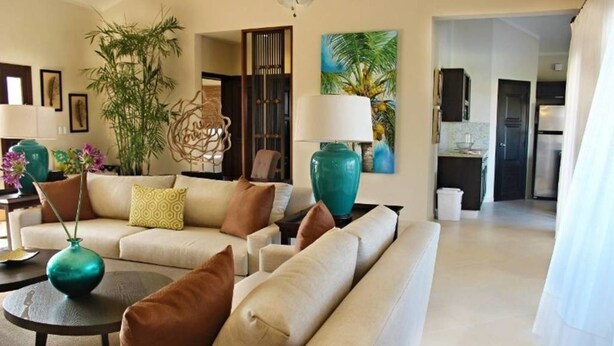 Un salón amplio con un sofá beige, una mesa de centro y jarrones decorativos. Hay una planta grande y un cuadro en la pared.