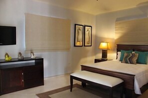 Una habitación de hotel con cama, mesita de noche con lámpara, un banco, cómoda y televisor en la pared.