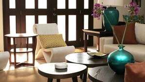 Un salón con un sofá, dos mesitas auxiliares, una silla y un ventanal con persianas de madera oscura.