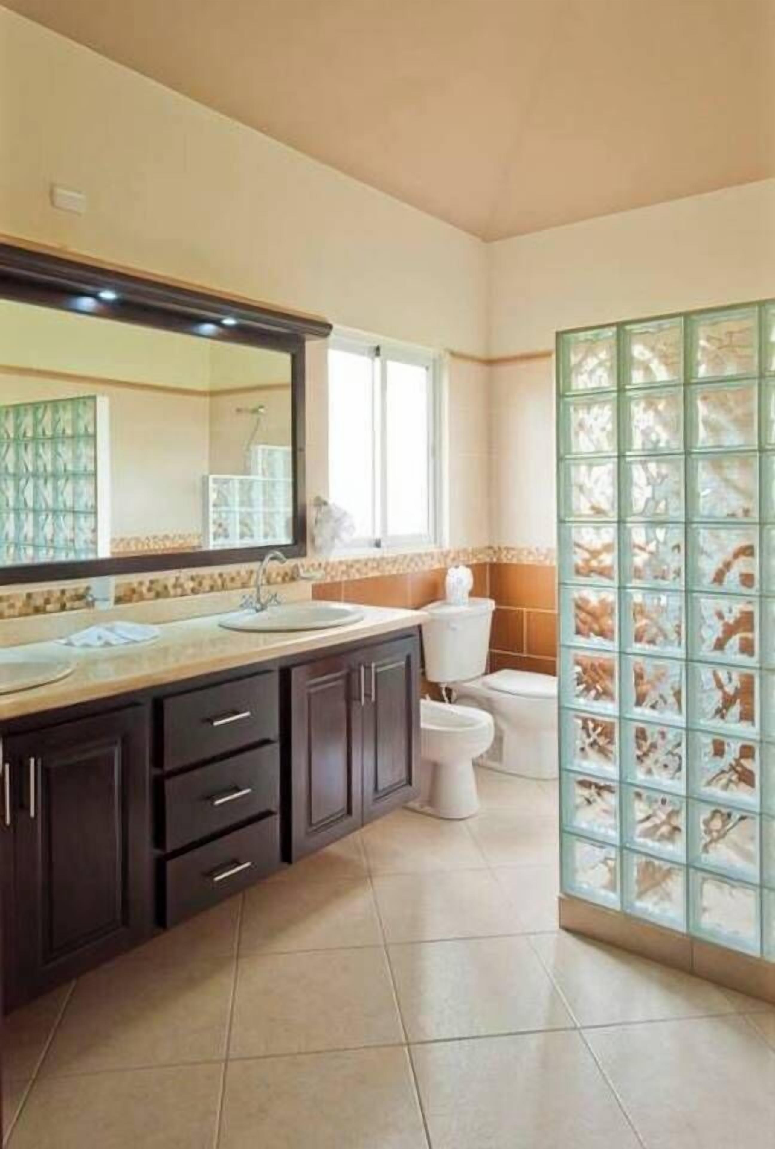 A bathroom with a large mirror, two sinks, a toilet, and a glass partition.