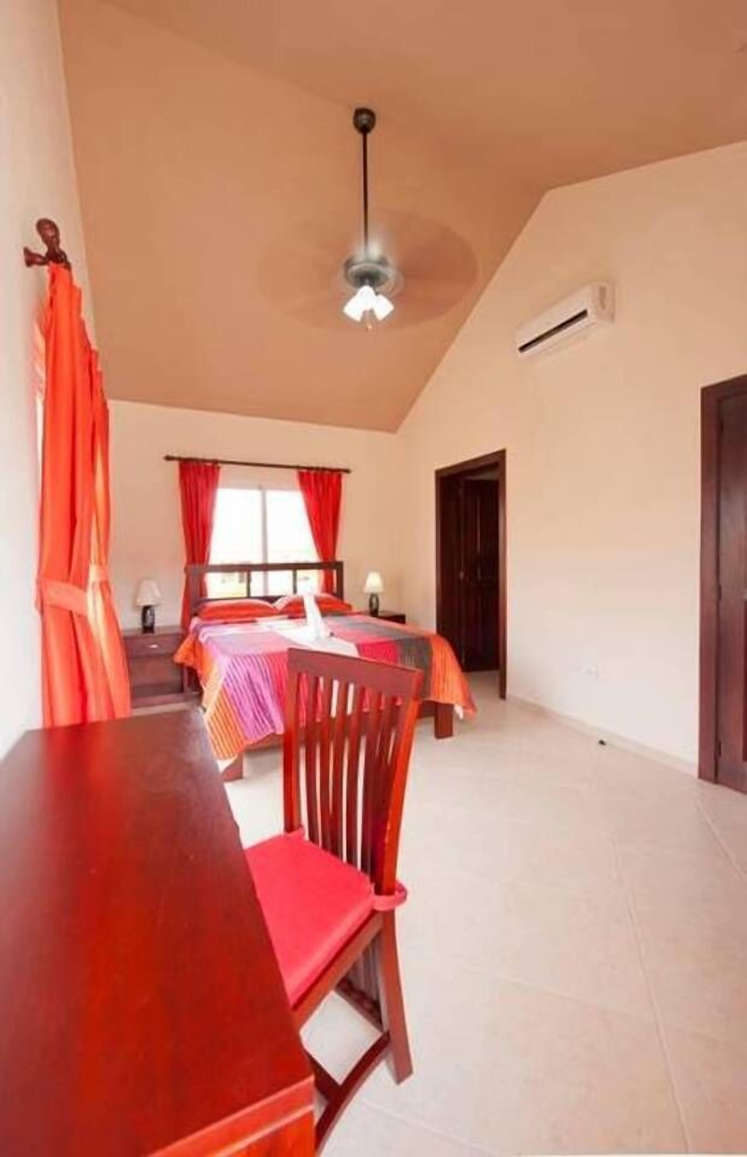A bedroom with a bed, a desk, and a chair. There is a ceiling fan, an air conditioning unit, and a window with curtains.