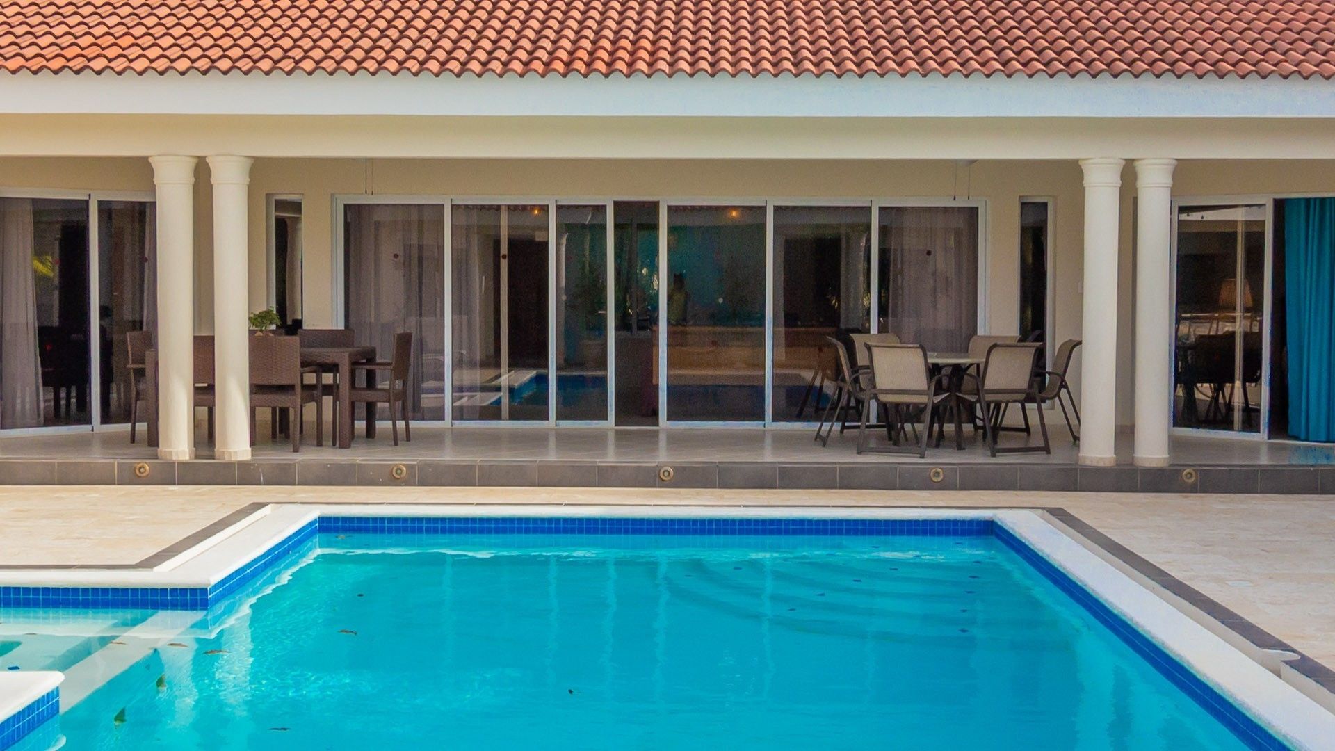 A swimming pool with a tiled deck, a covered patio area with chairs and tables, and a building with a tiled roof.