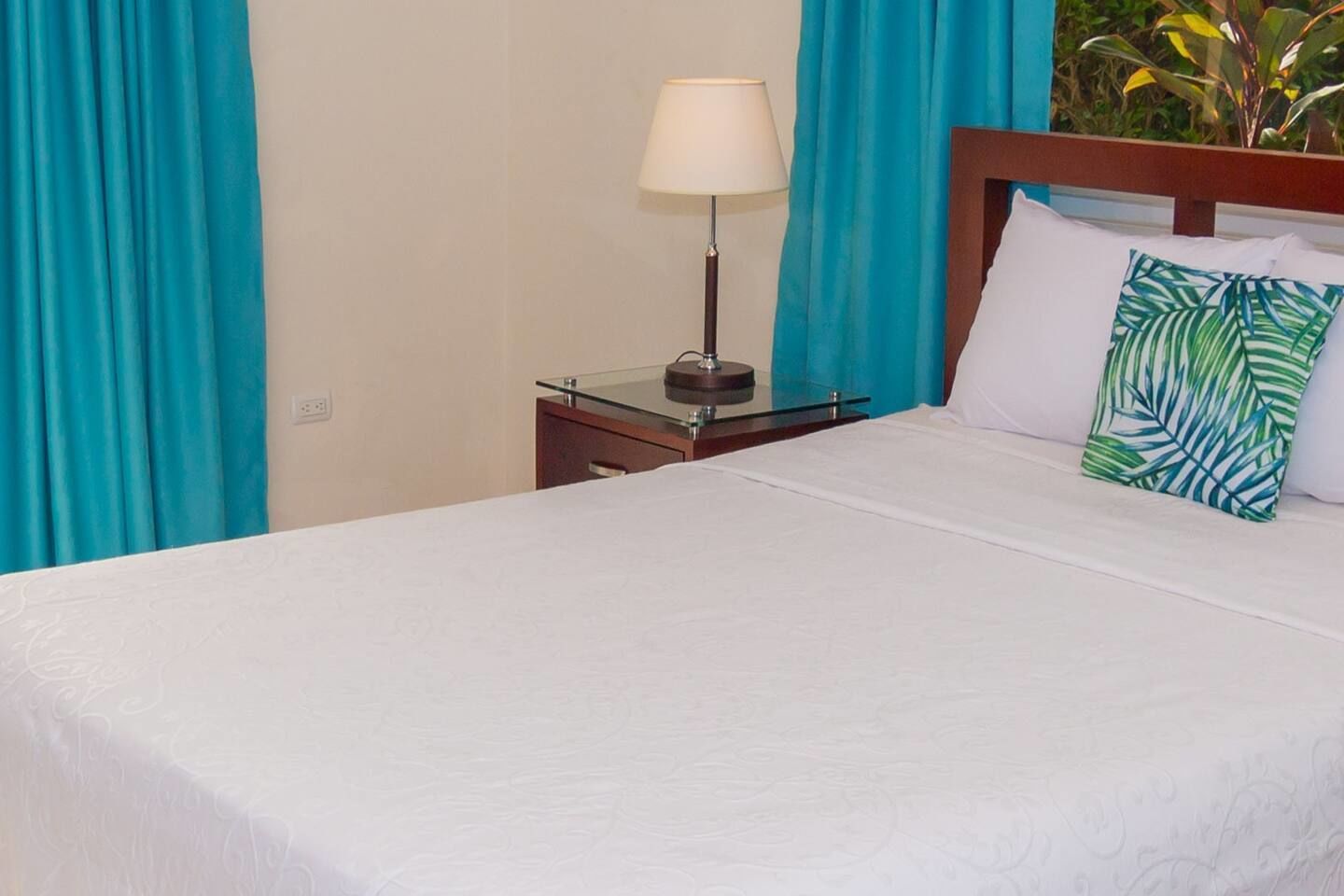 A neatly made bed with white bedding, a bedside lamp, and a tropical-themed pillow.