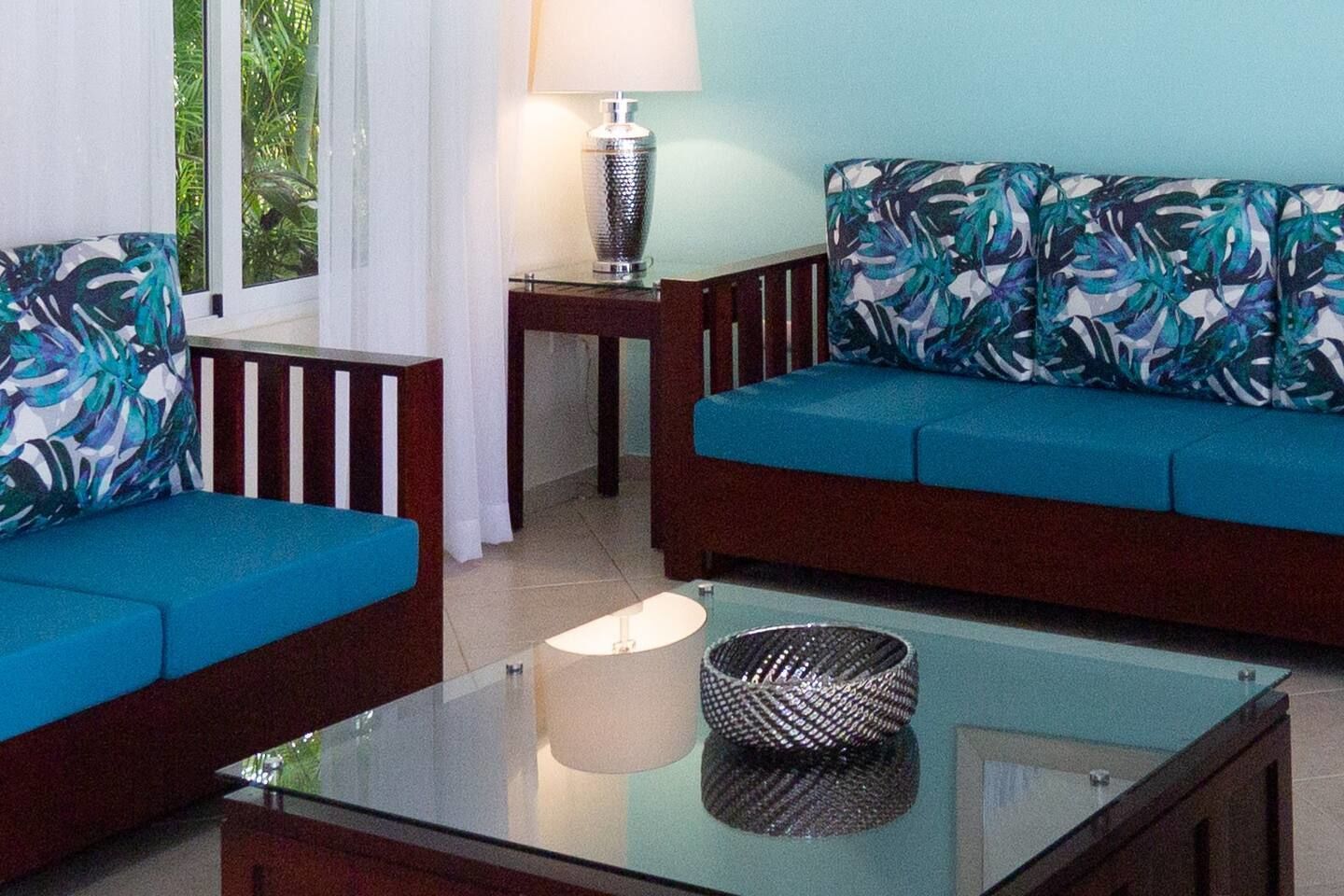 A living room with a glass coffee table, two sofas with patterned cushions, a small side table with a lamp, and a view of greenery through a window.