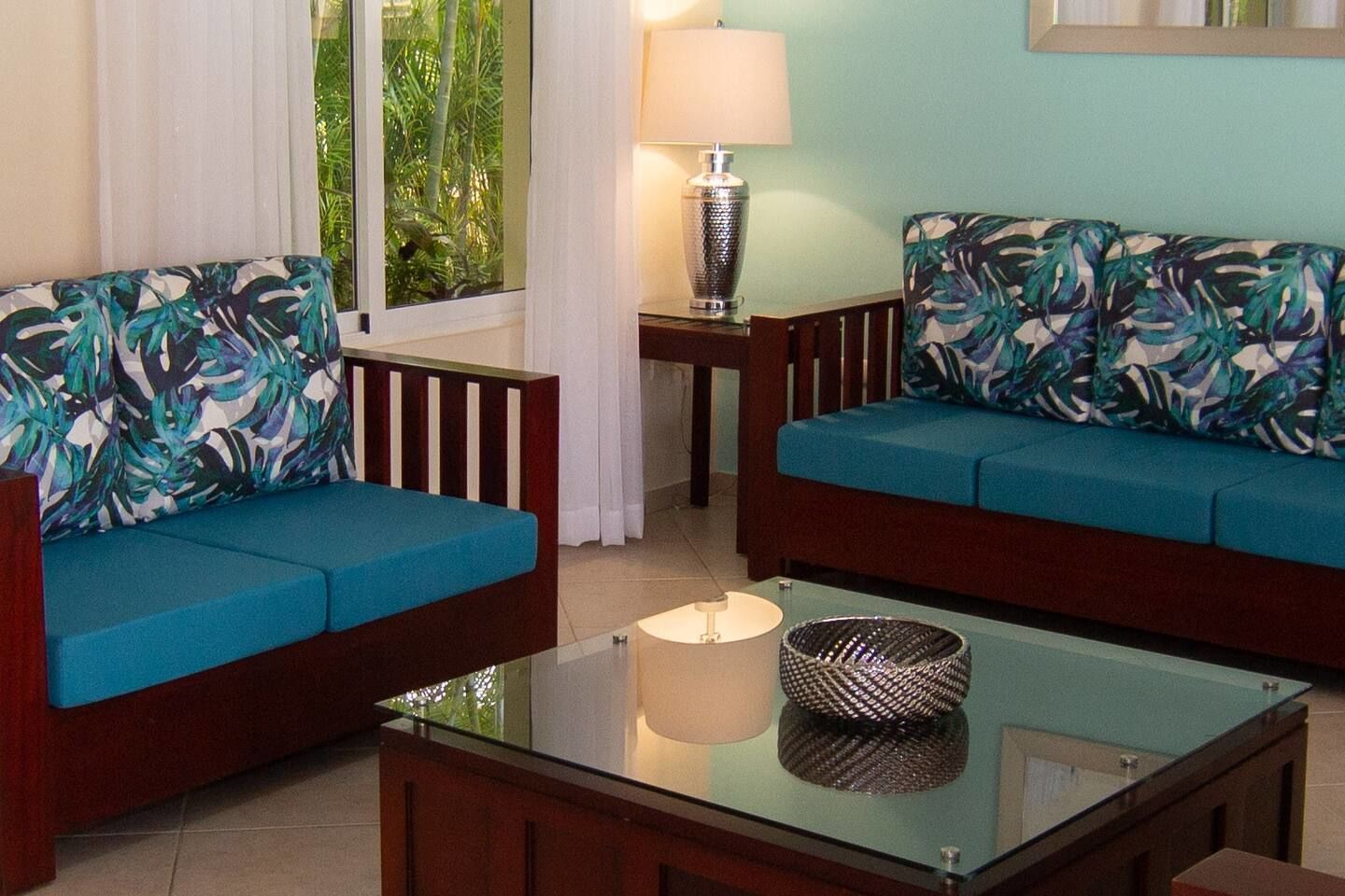 A living room with two blue sofas, a glass coffee table, a lamp, and a view of greenery through the window.