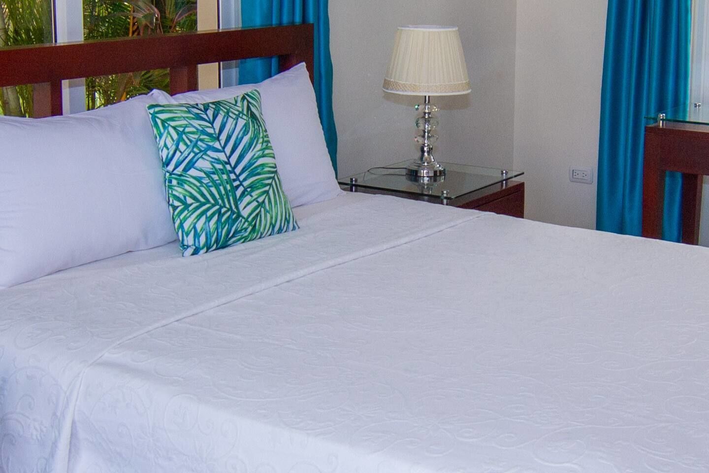 A neatly made bed with white linens, a green and blue patterned pillow, and a bedside table with a lamp.
