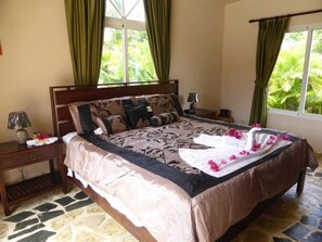 A bedroom with a large bed, green curtains, a nightstand, and a window with a view of greenery.