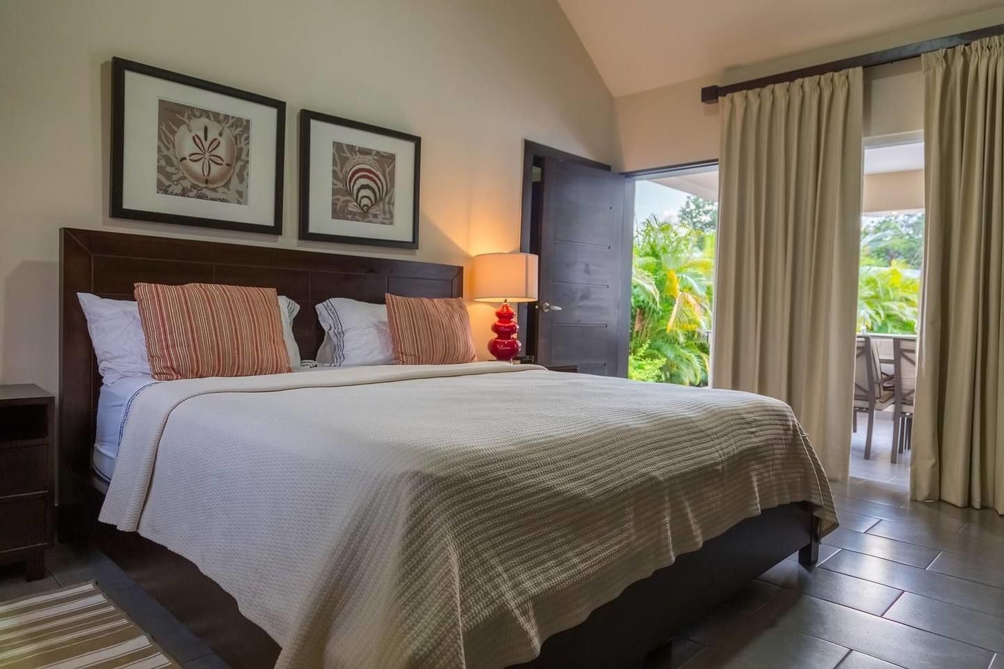 Une chambre d’hôtel avec un grand lit, deux œuvres d’art encadrées au-dessus, une table de chevet, une lampe et une vue sur l’extérieur grâce à une porte coulissante.