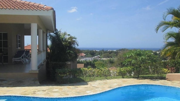 A swimming pool with a view of the ocean, a covered patio with chairs, and lush greenery. - Hilltop Ocean View Villa w/ Large Pool Walking to Strip and Sosua Beach (Sosúa)