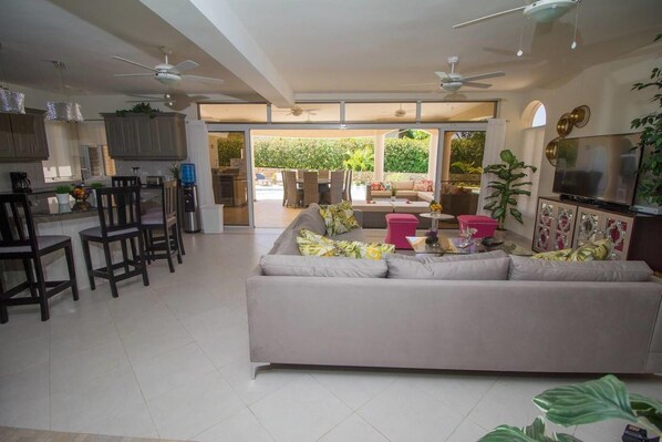 A spacious living area with a sofa, chairs, and a coffee table. There is a kitchen area with cabinets and a dining table. The room has large windows and a ceiling fan.
