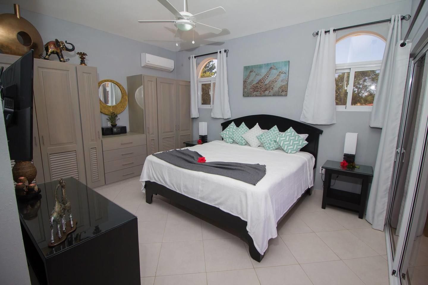 Une chambre à coucher comprenant un lit, un ventilateur de plafond, une commode, un miroir et des œuvres d’art accrochées aux murs.