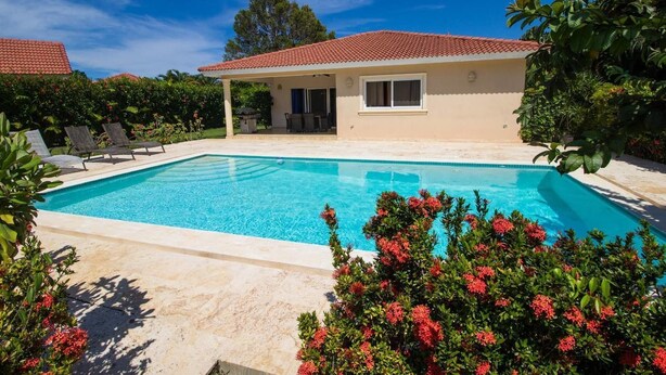 A rectangular swimming pool with a tiled deck, surrounded by lush greenery and red flowers. - Sunny Days and Tropical Nights Await you (Sosúa)