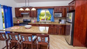 A kitchen with granite countertops, wooden cabinets, a central island with chairs, a stove, a microwave, and a refrigerator. - Sunny Days and Tropical Nights Await you (Sosúa)