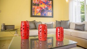 Un salon avec une table basse en verre, trois vases rouges, un canapé avec des coussins gris et un tableau abstrait aux couleurs vives accroché au mur.