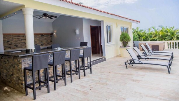 Un espace bar extérieur doté d’un comptoir en pierre et de chaises en osier, d’un ventilateur de plafond et d’un coin salon au bord de la piscine avec des chaises longues.