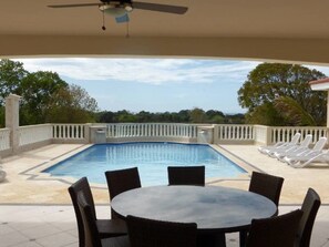 A pool area with a table and chairs, a ceiling fan, and a view of trees and a building in the background.
