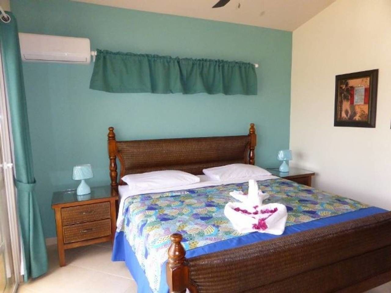 Un dormitorio con cama de madera, paredes color turquesa, una ventana con cortina, una mesita de noche con lámpara y un cuadro en la pared.