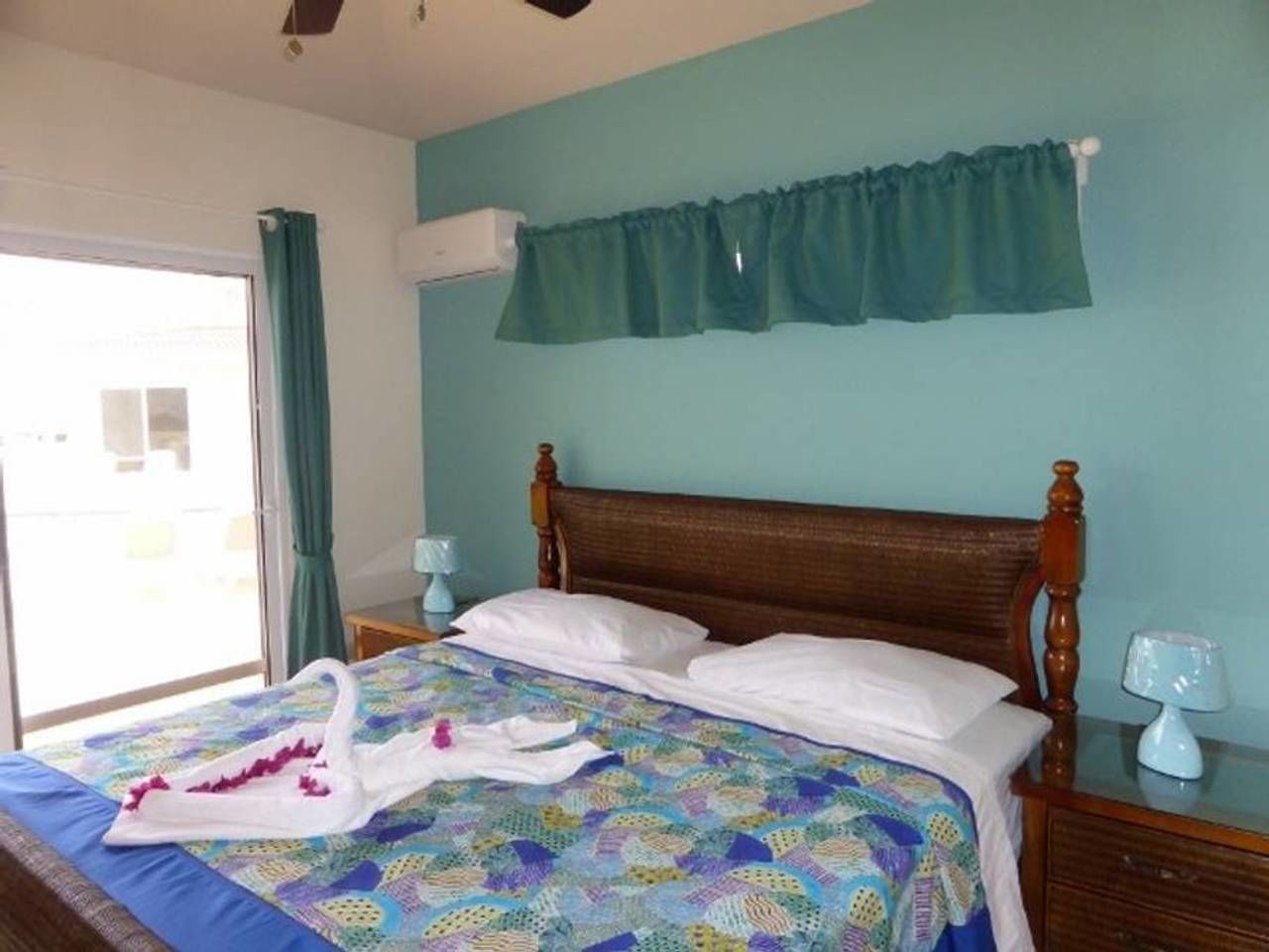 Een slaapkamer met een houten bed, een bed met een patroon, een blauwgroen gordijn en een raam met uitzicht.