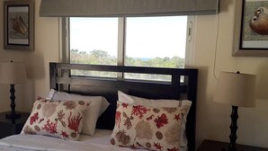 Cama com roupa de cama branca e almofadas decorativas, janela com vista para o verde e mesa de cabeceira com luminária.