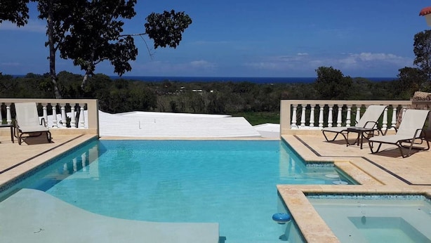 Uma piscina infinita retangular com vista para o oceano, rodeada por espreguiçadeiras e um guarda-corpo branco.