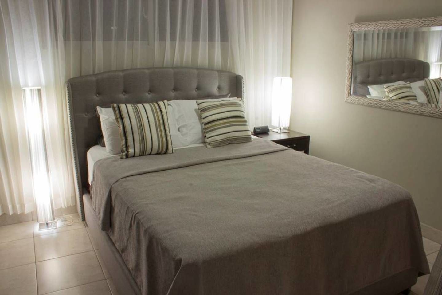 A neatly made bed with a grey headboard and striped pillows, a bedside table with a lamp, and a mirror on the wall.