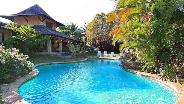 A swimming pool with a stone border, surrounded by lush greenery and a house with a tiled roof. - Villa Las Palmas Caribbean Family Vacation Rental (Cabarete)
