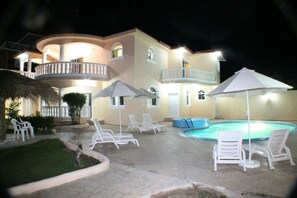A two-story building with a swimming pool, white lounge chairs, and umbrellas.