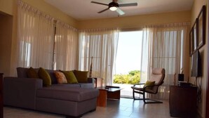 A living room with a sofa, armchair, and coffee table. There is a ceiling fan, large windows with curtains, and a TV on the wall. - Ocean View 2 Bedroom Villa Newly Build in Gated Community (Sosúa)