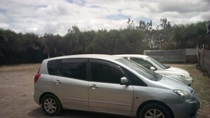 Un coche utilitario plateado estacionado sobre una superficie de tierra, con un área verde y árboles al fondo.