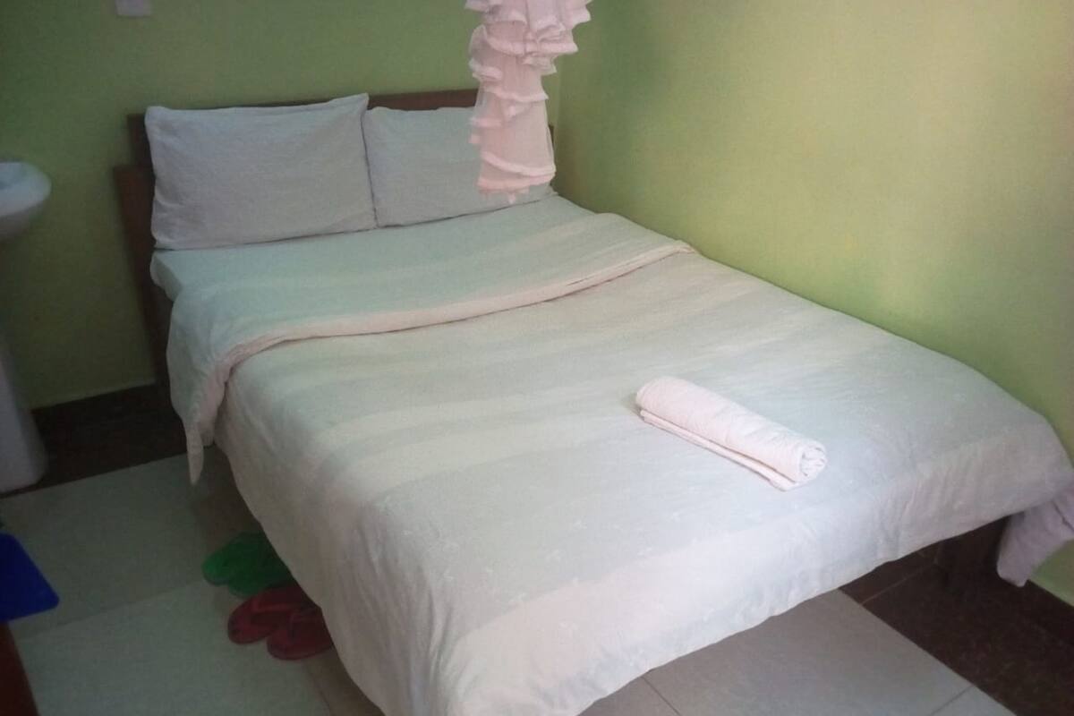 A single bed with a white bedspread and a rolled towel on it. There is a pillow on the bed, and a small table with a white cloth is visible to the left.