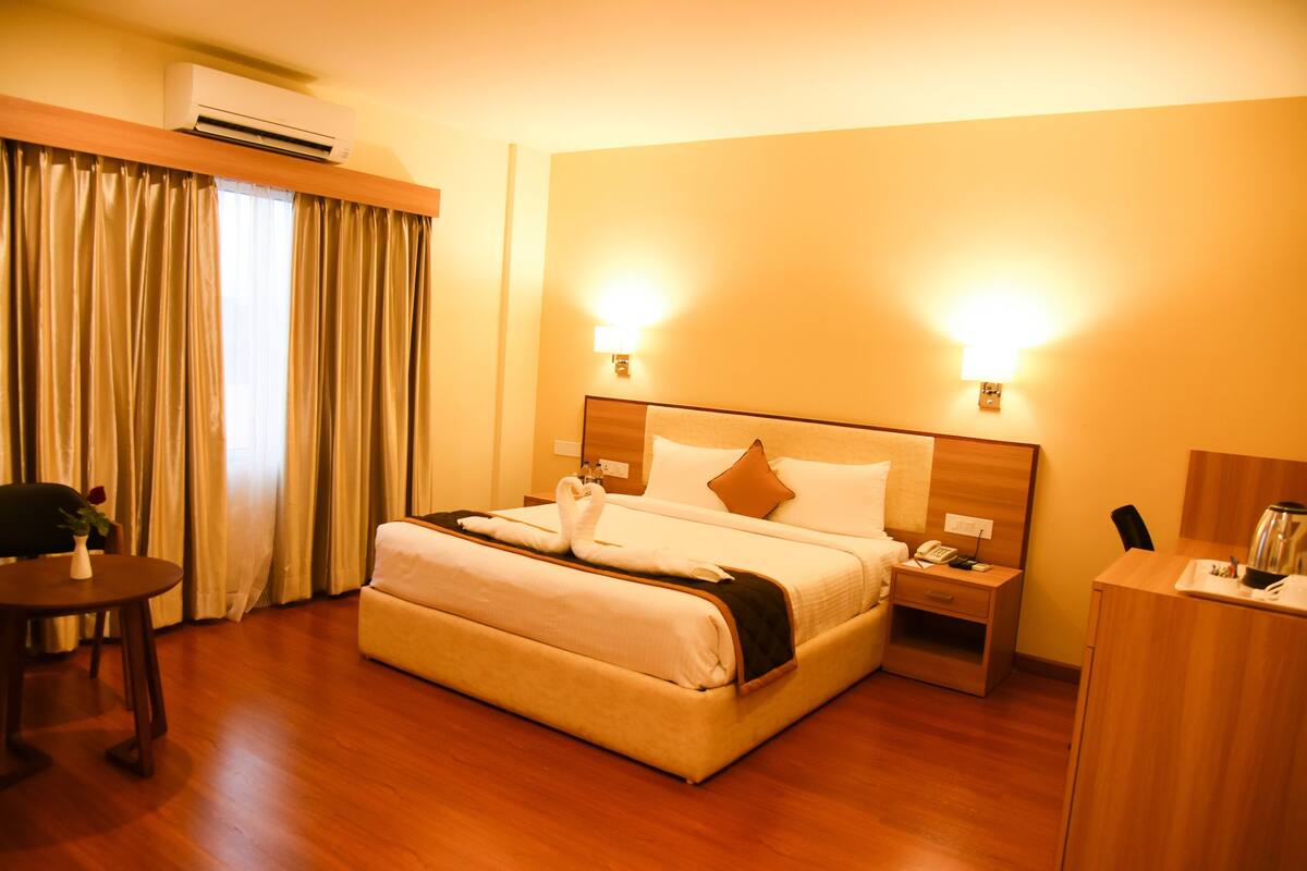 A hotel room with a large bed, bedside tables, an air conditioning unit, and a desk with a chair.