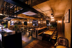 A restaurant with wooden tables and benches, exposed brick walls, and hanging light fixtures. - Hotel RK Gardenia (Anekal)