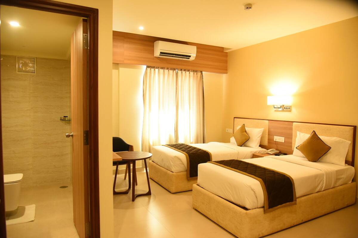 A hotel room with two beds, a small table, a chair, an air conditioning unit, and a shower area.
