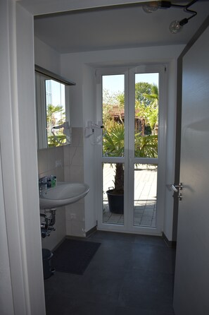 Une salle de bain avec un lavabo blanc, un miroir et une porte donnant sur un espace extérieur agrémenté d’une plante en pot.