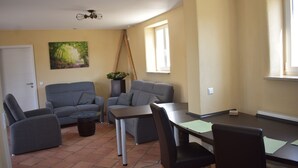 En stue med en grå sofa, et lille bord og en stol. Der er et billede på væggen og et vindue med persienner.