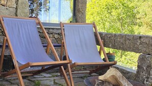 Two wooden lounge chairs with white cushions, a metal fire pit with wood and kindling, and a stone wall in the background.