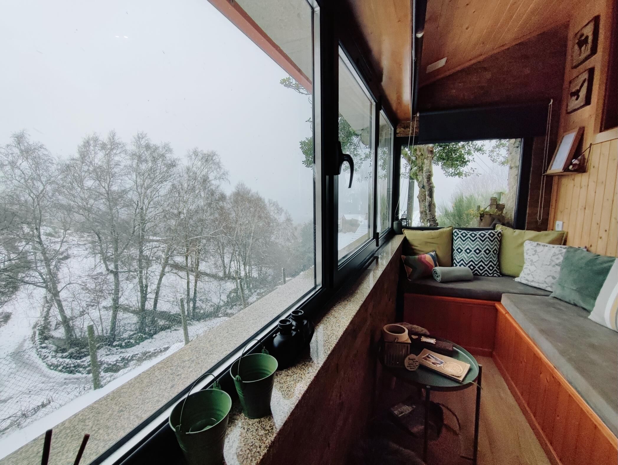 Een gezellig cabin interieur met een houten bank en kussens, een kleine tafel met spullen, en een raam met uitzicht op een besneeerd landschap.