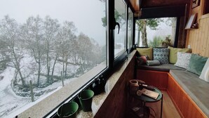 A cozy cabin interior with a wooden bench and cushions, a small table with items, and a window overlooking a snowy landscape.