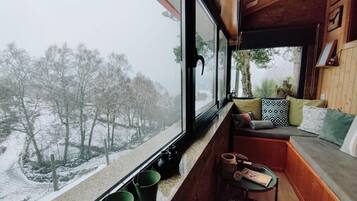 Een gezellig cabin interieur met een houten bank en kussens, een kleine tafel met spullen, en een raam met uitzicht op een besneeerd landschap.