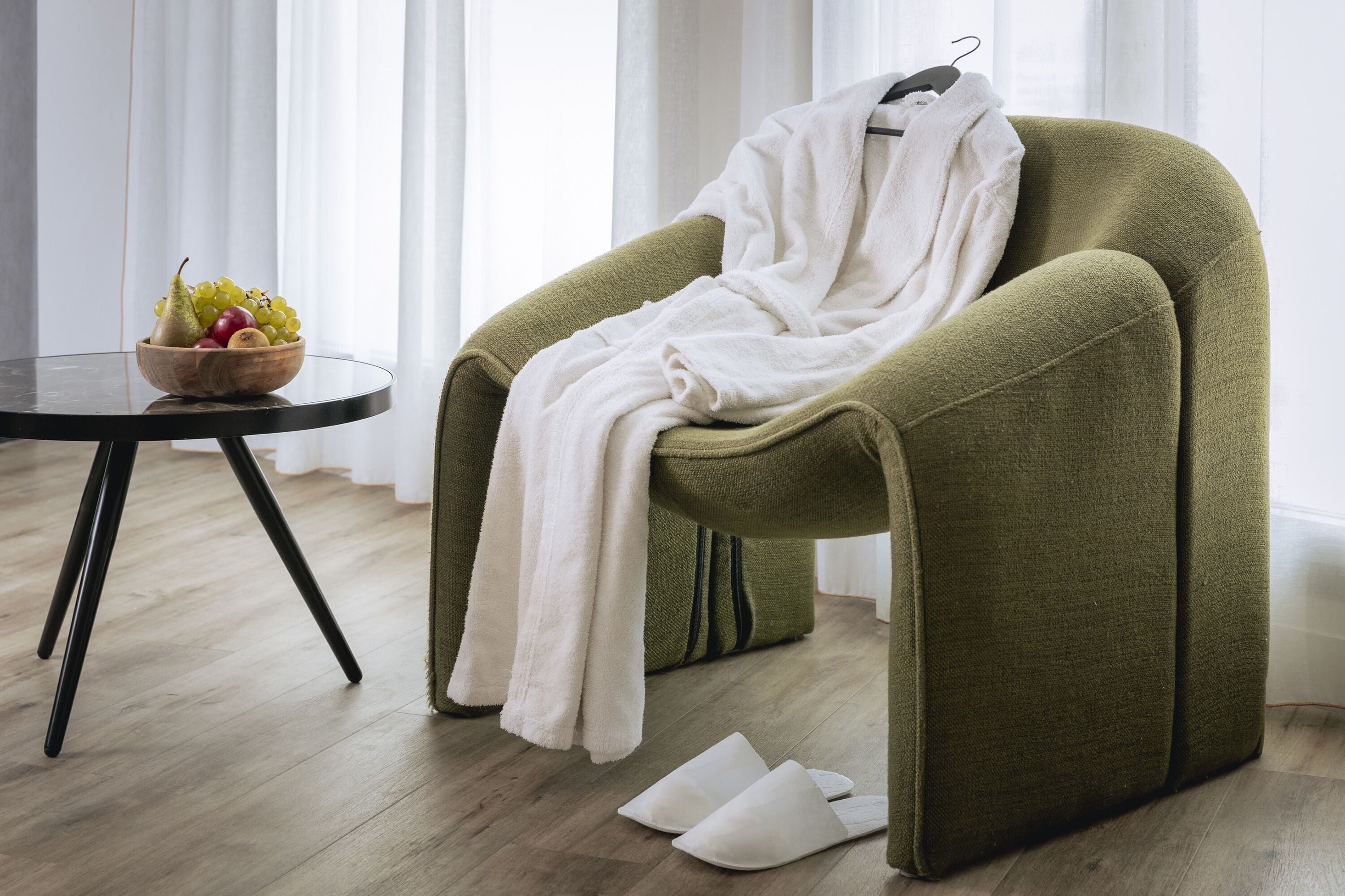 A room with a green armchair, a white bathrobe, a bowl of fruit, and a pair of slippers on the floor.