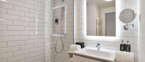 Un baño moderno con azulejos blancos, ducha con mampara de vidrio, lavamanos con espejo y dos botellas de champú.