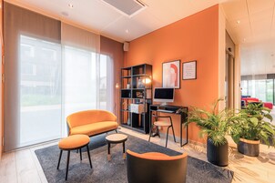 Una habitación de hotel moderna con un ventanal grande, un sofá naranja, un escritorio con una computadora y dos plantas en macetas.