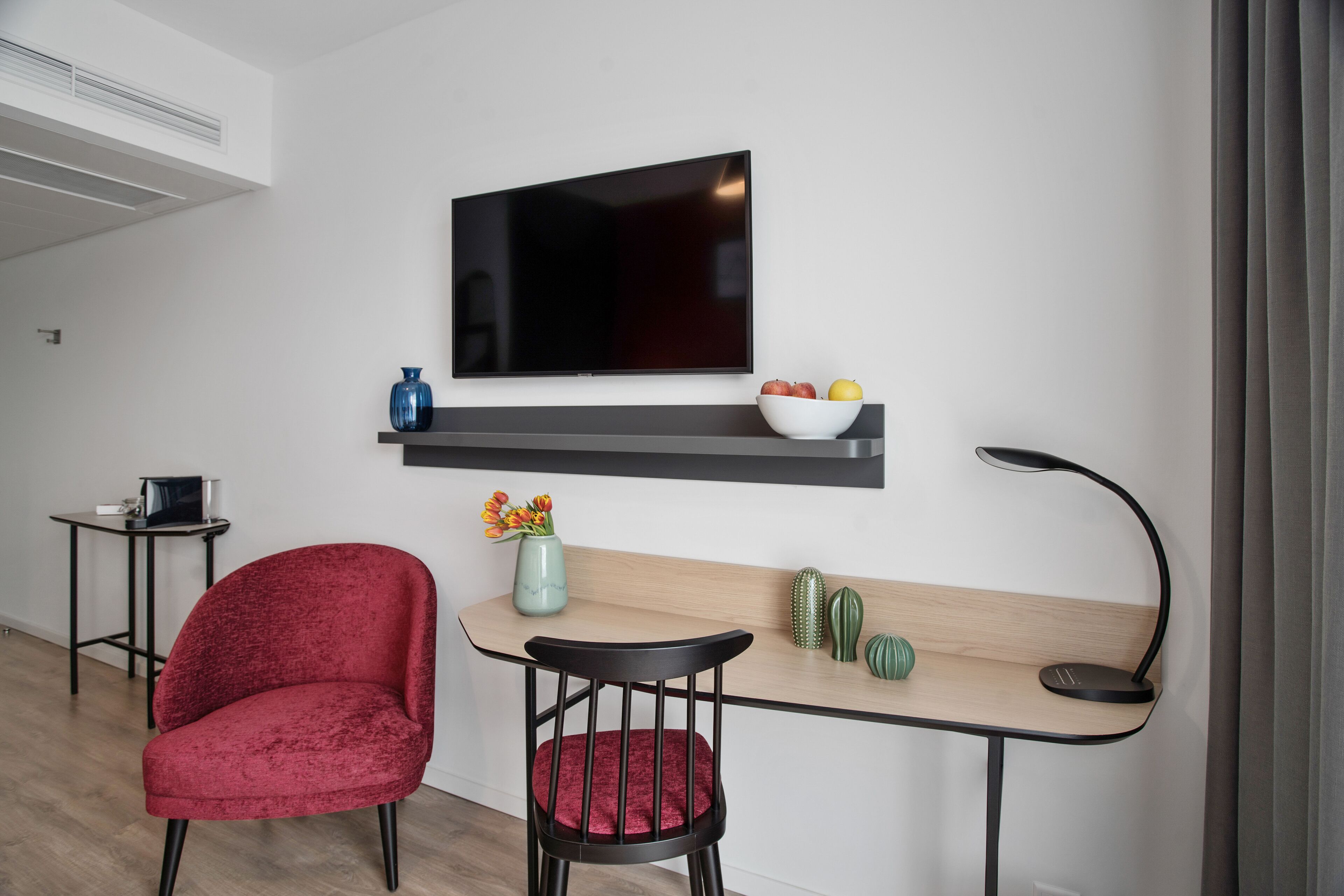 Ett modernt vardagsrum med en platt-TV, ett skrivbord i trä, en röd stol, ett litet bord och en kaktus på väggen.