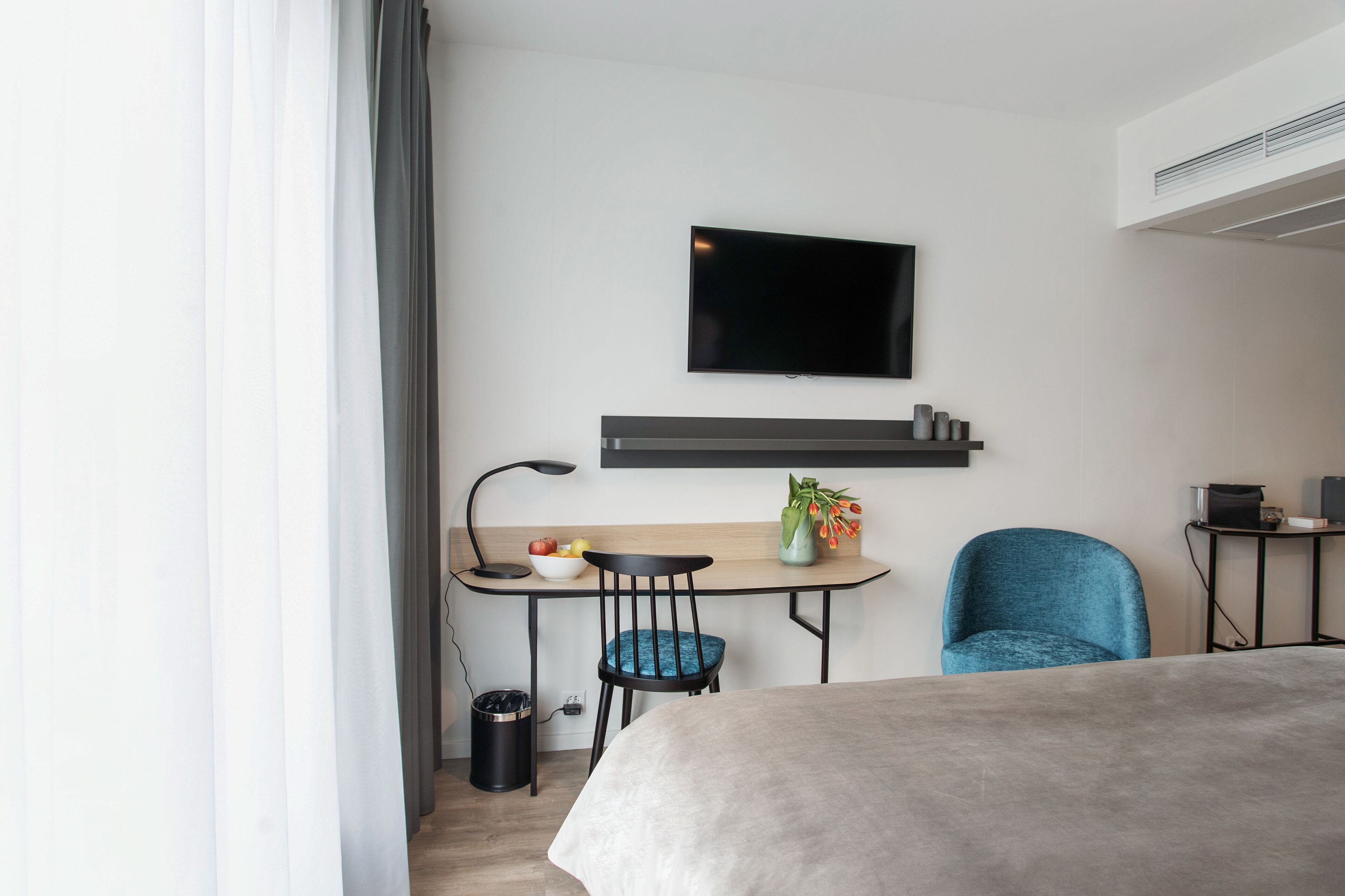 A modern hotel room with a bed, a desk with a chair, a television mounted on the wall, and a small table with a vase of flowers.