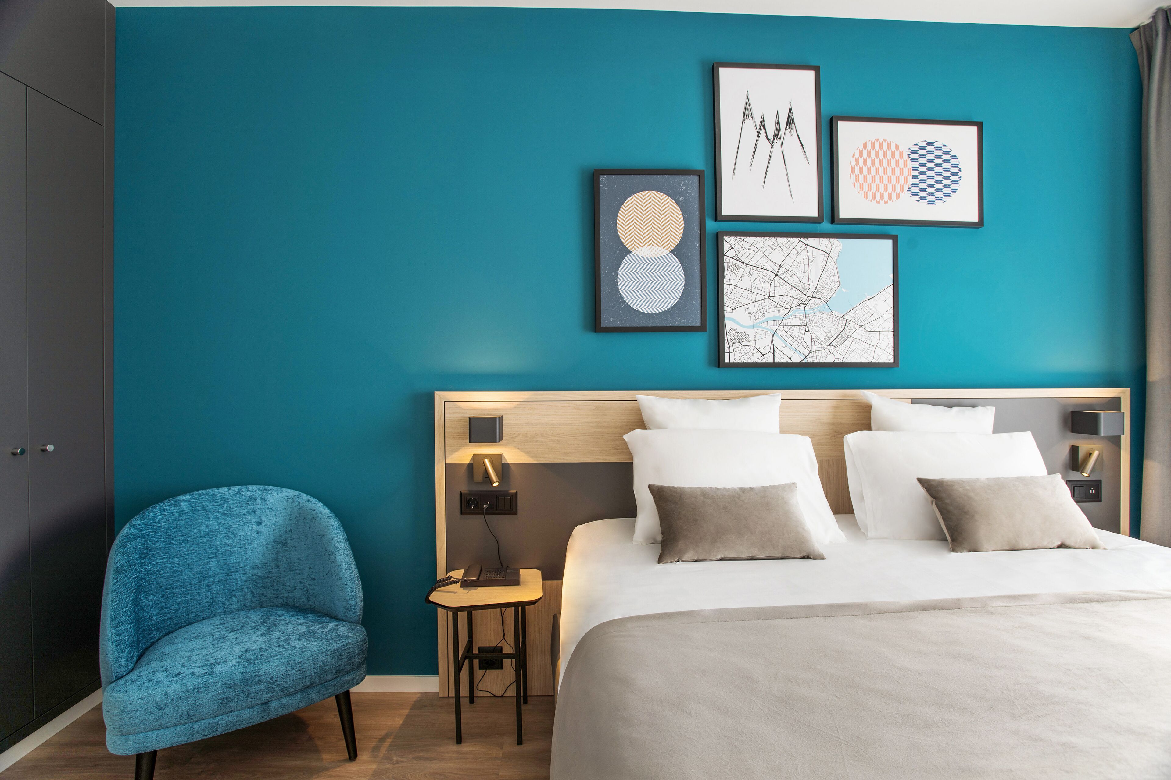 Habitación de hotel moderna con paredes verde azulado, una cama grande, un sillón azul y cuadros en la pared.