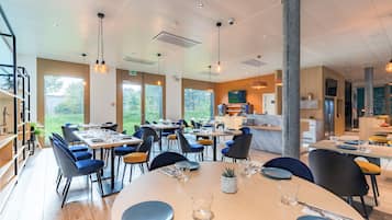 Un espace repas moderne doté de plusieurs tables et chaises, de grandes fenêtres et d’une cuisine ouverte.