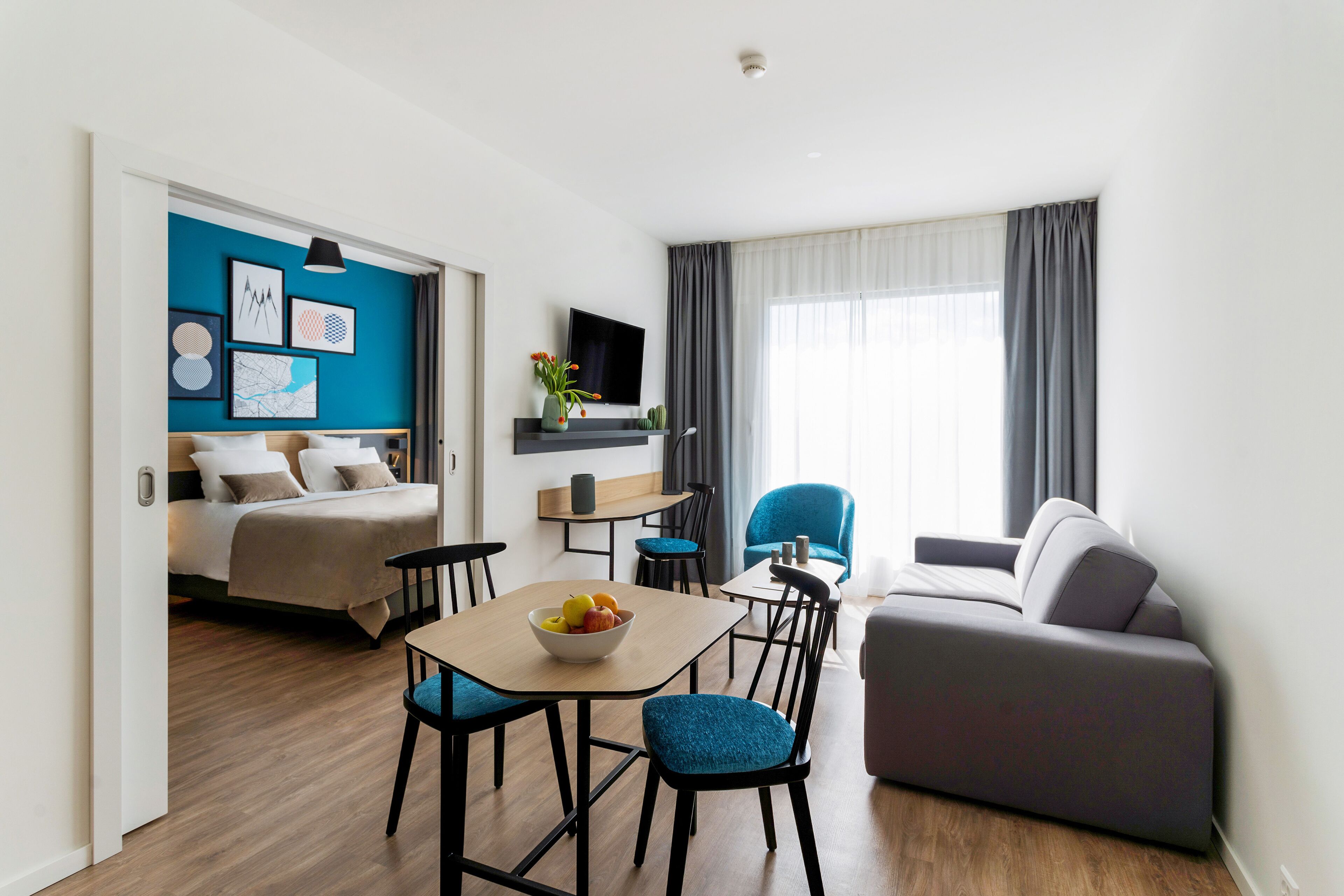 Une chambre d’hôtel moderne équipée d’un lit, d’un coin repas avec une table et des chaises, d’un canapé et d’un téléviseur fixé au mur.